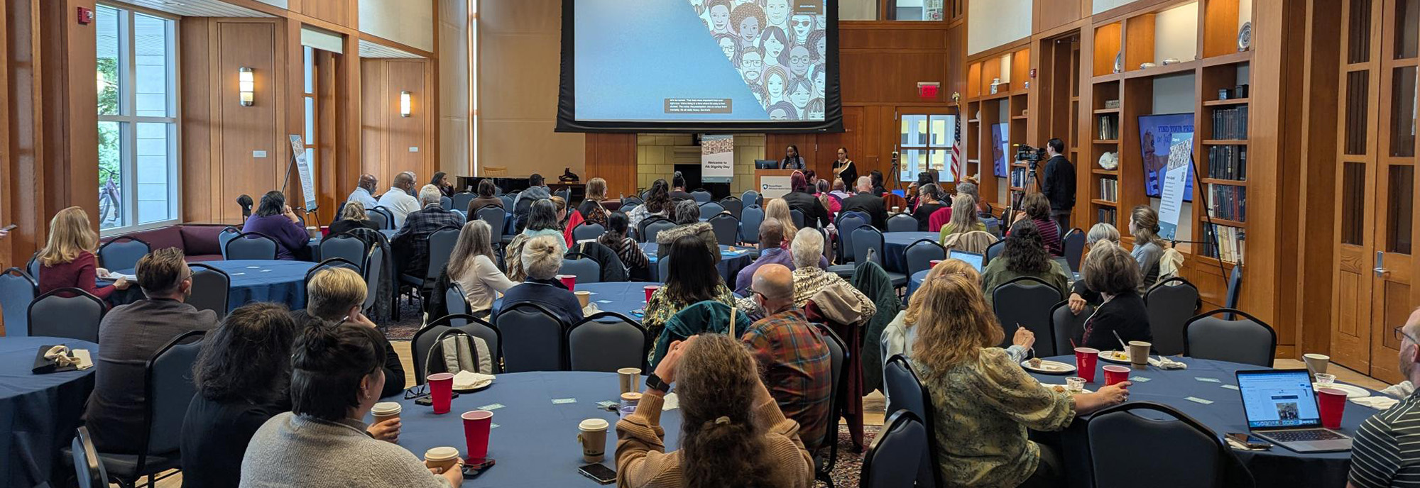 Conference attendees view a presentation on a large screen at the front of a bright, open room with high ceilings.