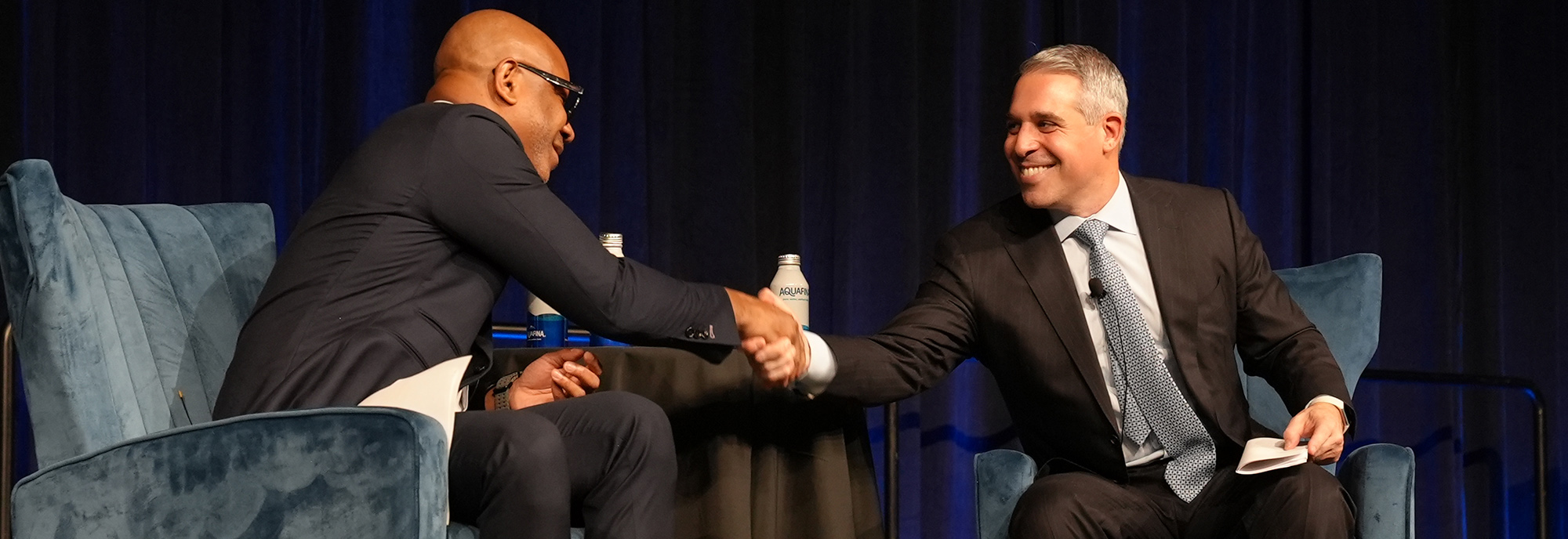 Two men shake hands on stage during a panel discussion at a large conference.